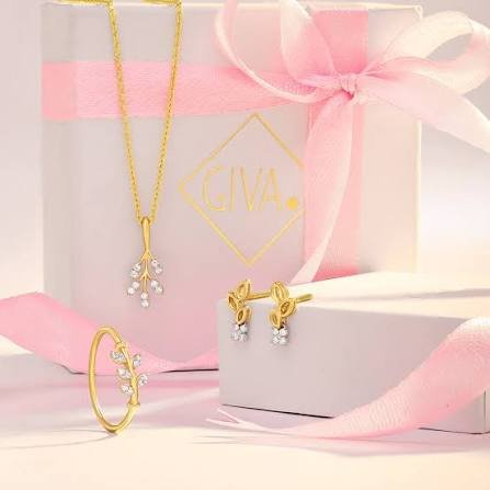 A GIVA gold-plated pendant chain, matching studs, and a delicate leaf-shaped ring arranged around a pastel gift box tied with a pink ribbon